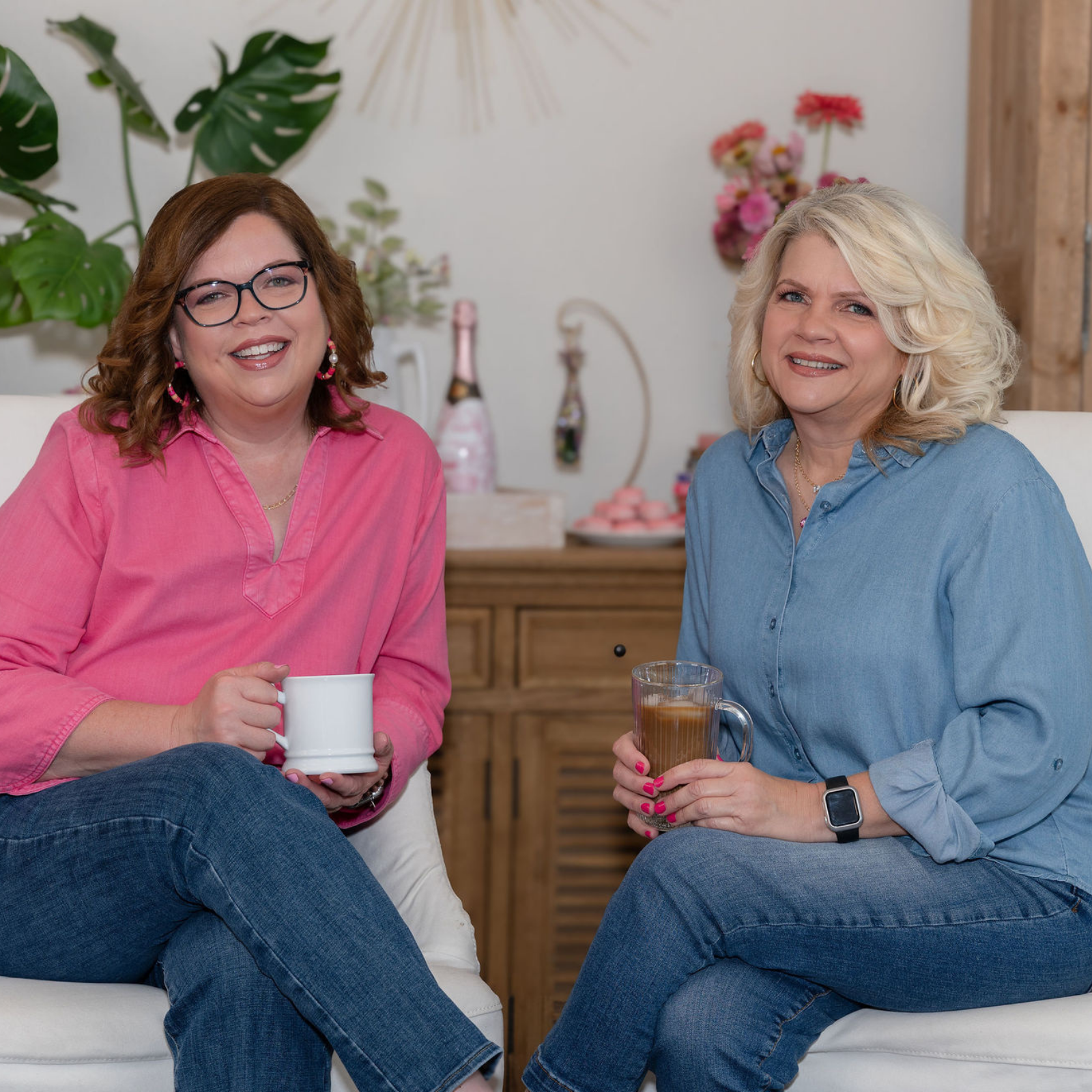 Two women sitting on white chairs in a bright, decorated room, holding mugs and glasses. The woman on the left wears a pink button-up shirt and blue jeans, while the woman on the right wears a light blue button-up shirt and jeans.