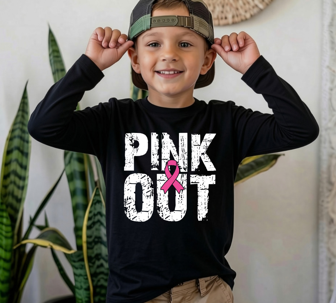 A smiling young boy wearing a black long-sleeve t-shirt with a 'PINK OUT' graphic and a camouflage baseball cap, posing indoors with his hands on his ears.