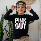 A smiling young boy wearing a black long-sleeve t-shirt with a 'PINK OUT' graphic and a camouflage baseball cap, posing indoors with his hands on his ears.