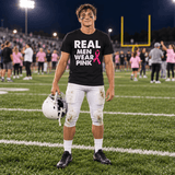 A football player on a field at night wearing a black graphic T-shirt that reads 'REAL MEN WEAR PINK' with a pink ribbon, white pants, and holding a helmet.
