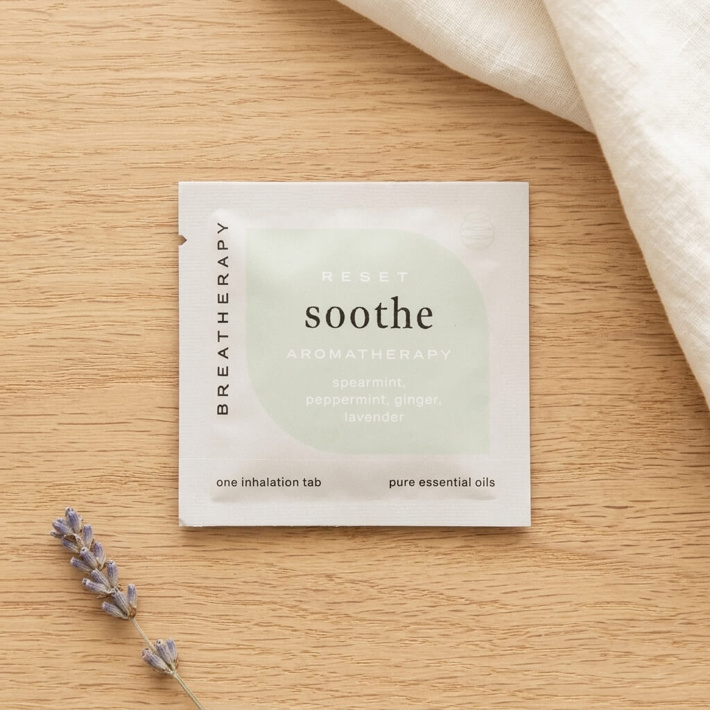 A 'soothe' aromatherapy inhalation tab packet lies on a light wood surface, styled with a sprig of lavender and a folded cream fabric in the corner.
