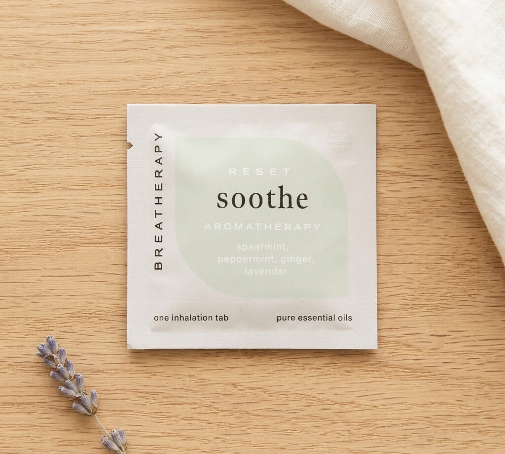 A 'soothe' aromatherapy inhalation tab packet lies on a light wood surface, styled with a sprig of lavender and a folded cream fabric in the corner.