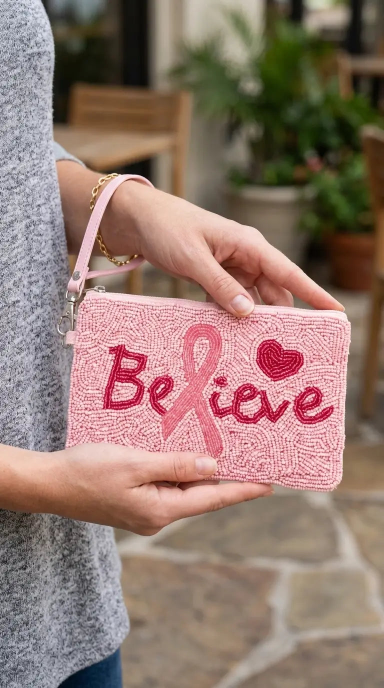 A person holding a pink beaded wristlet clutch with 'Believe' and a pink ribbon embroidered on the front, set against an outdoor patio background.