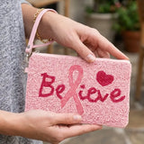 A person holding a pink beaded wristlet clutch with 'Believe' and a pink ribbon embroidered on the front, set against an outdoor patio background.