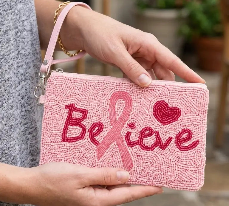 A person holding a pink beaded wristlet clutch with 'Believe' and a pink ribbon embroidered on the front, set against an outdoor patio background.