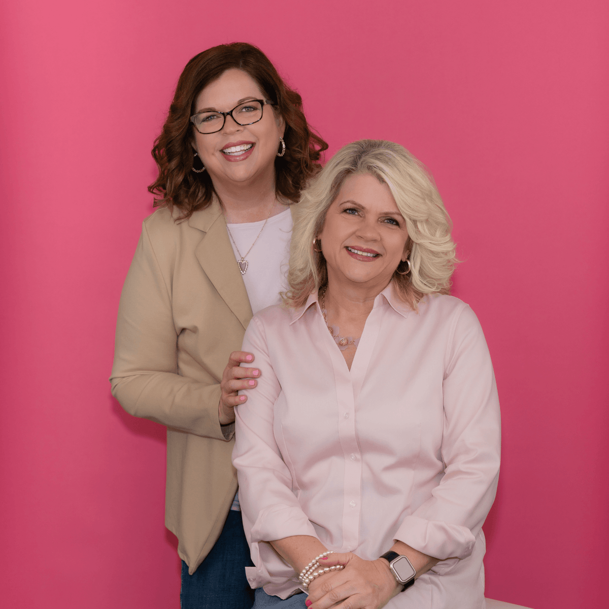Two women posing together in front of a solid pink background. The woman on the left has dark brown hair, wears glasses, a beige blazer, and a white top. The woman on the right has platinum blonde hair and wears a pink top with her hands crossed.