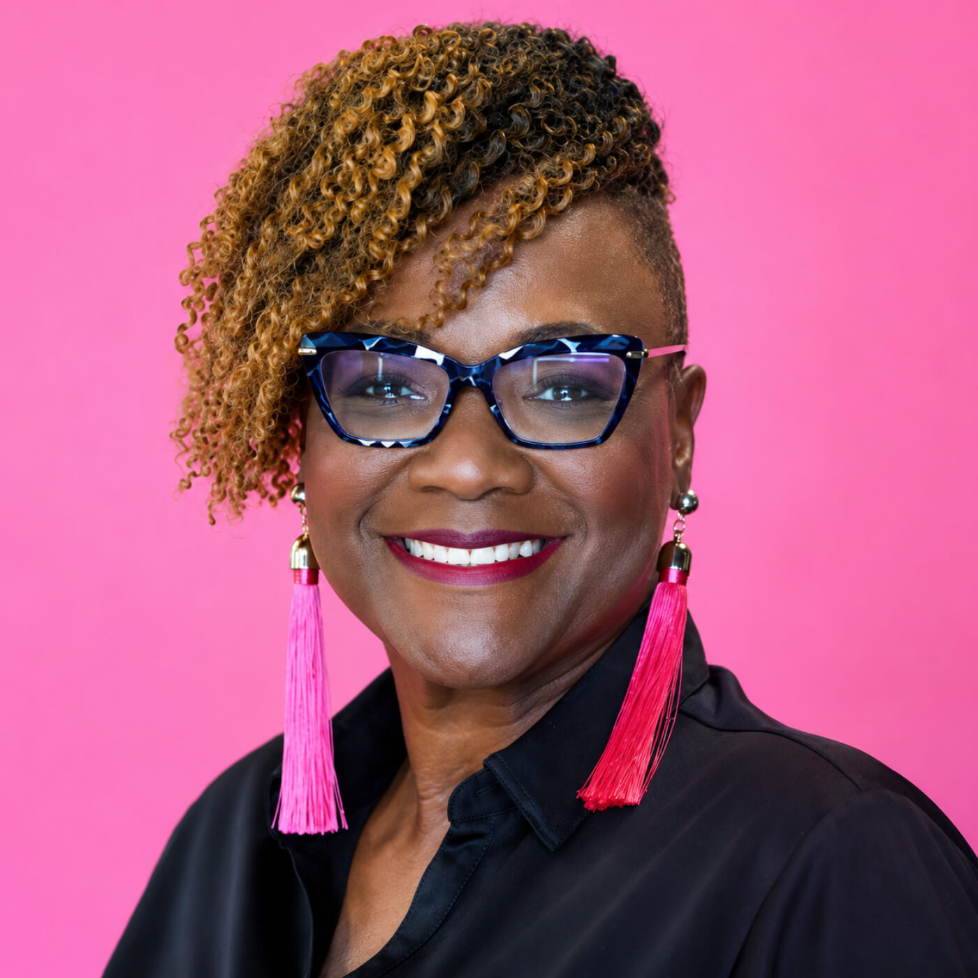 Hilda Marcel, a smiling woman with short curly hair, wearing blue-framed glasses and bright pink tassel earrings, against a solid pink background.