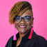 Hilda Marcel, a smiling woman with short curly hair, wearing blue-framed glasses and bright pink tassel earrings, against a solid pink background.