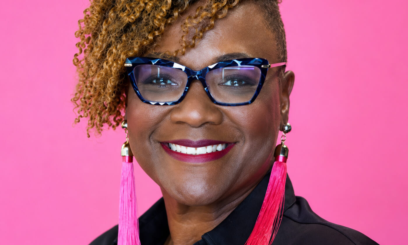 Hilda Marcel, a smiling woman with short curly hair, wearing blue-framed glasses and bright pink tassel earrings, against a solid pink background.