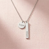 The Filled with Hope pendant necklace in silver stainless steel, resting on a soft pink background.
