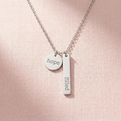 The Filled with Hope pendant necklace in silver stainless steel, resting on a soft pink background.
