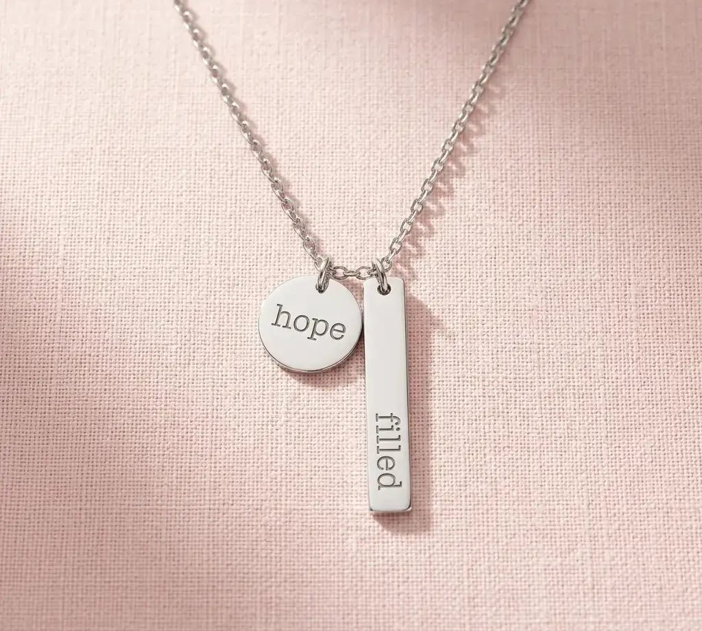The Filled with Hope pendant necklace in silver stainless steel, resting on a soft pink background.
