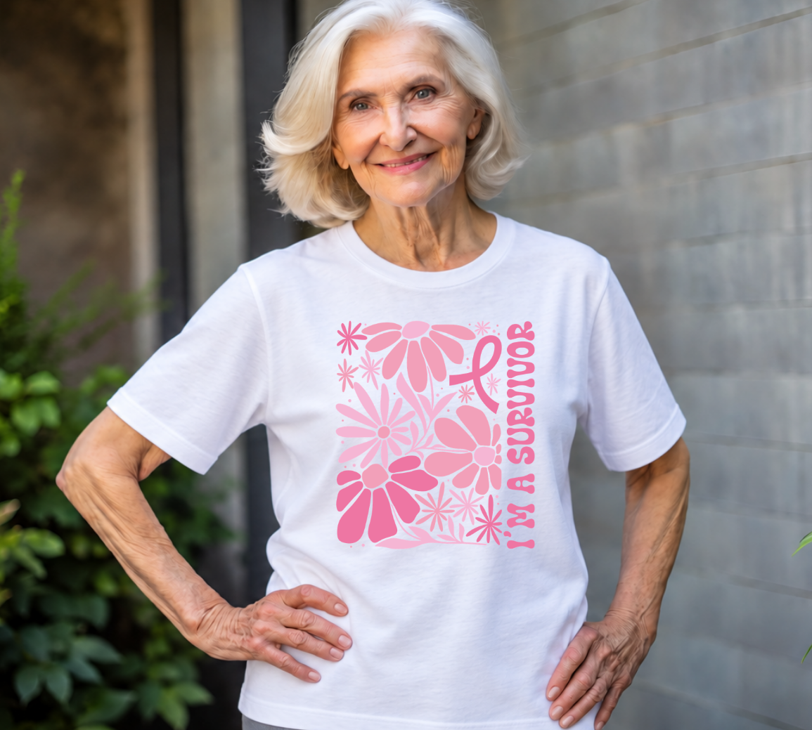 An older woman with short white hair stands outdoors wearing a white short-sleeve T-shirt with a pink floral graphic and the words 'I'M A SURVIVOR' on the front.