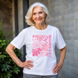An older woman with short white hair stands outdoors wearing a white short-sleeve T-shirt with a pink floral graphic and the words 'I'M A SURVIVOR' on the front.