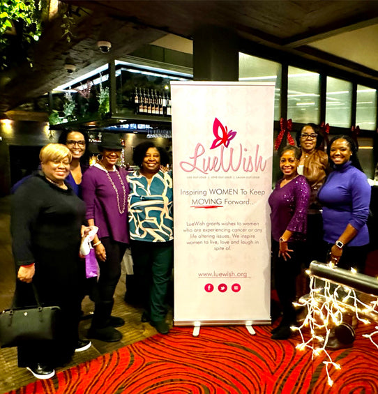 A group of seven women standing together in front of a white LueWish promotional banner on a red patterned carpet in an indoor event space.