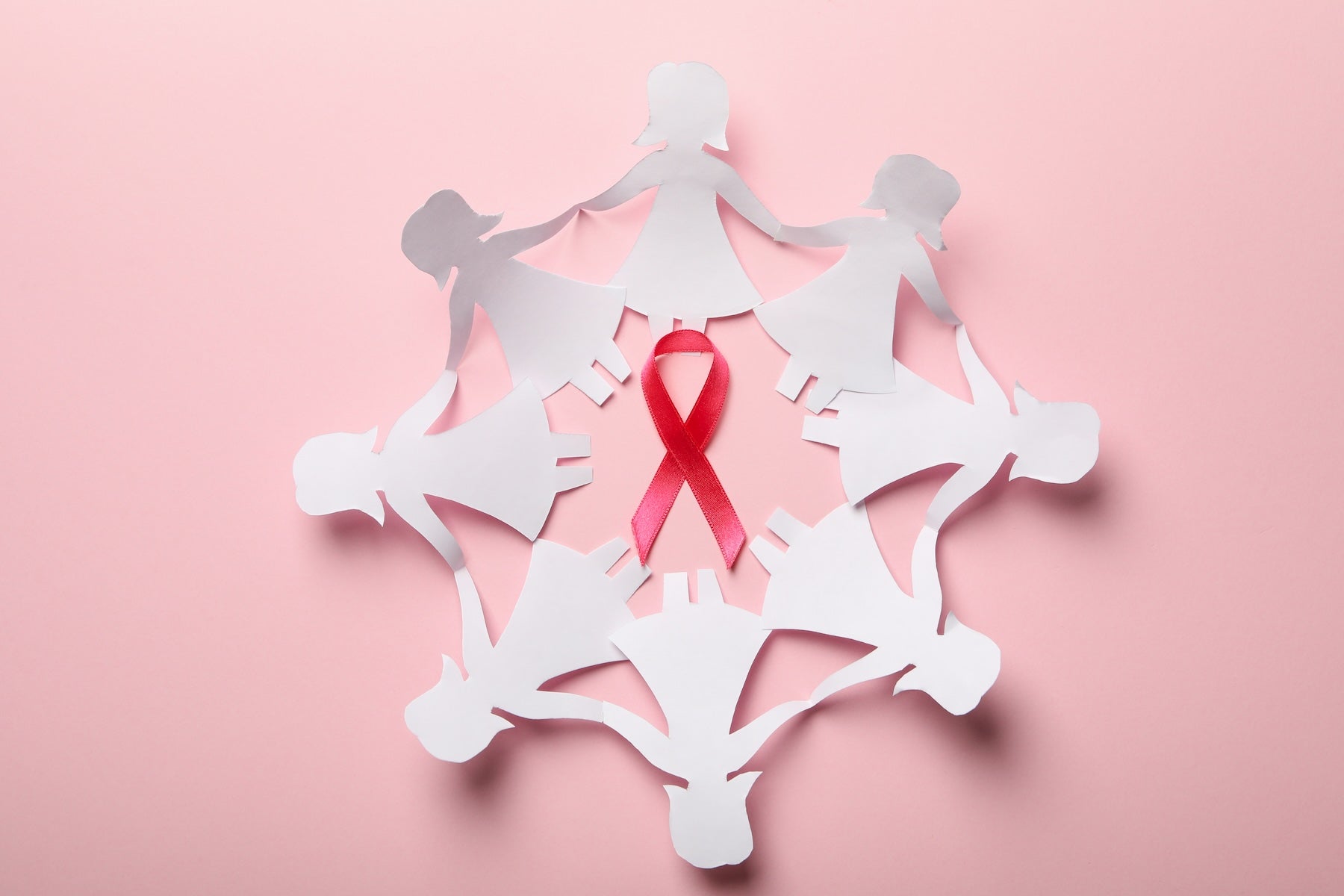 A pink awareness ribbon centered inside a circular arrangement of white paper cutout figures holding hands, set against a solid pink background.
