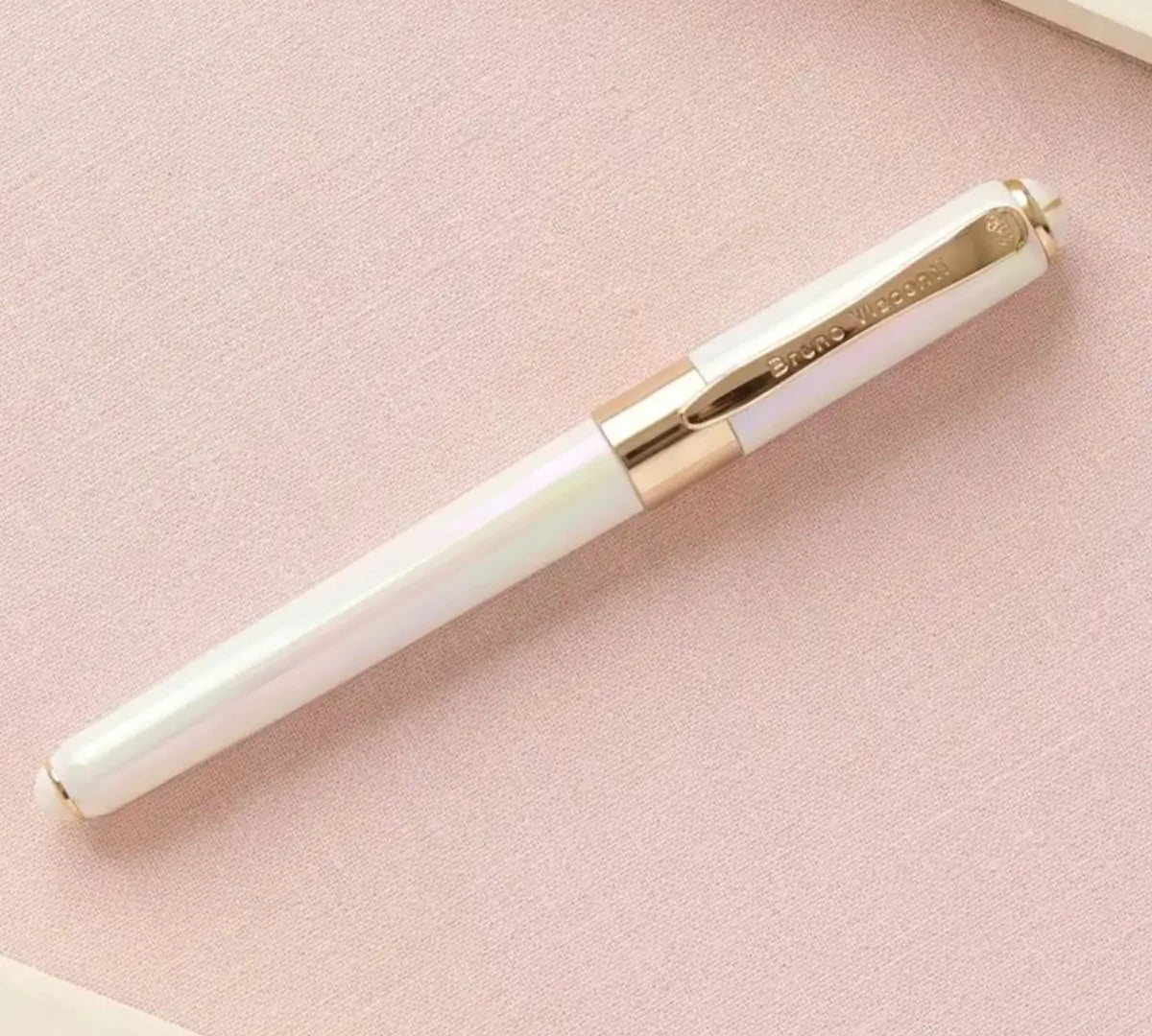 A white iridescent rollerball pen with gold trim and a gold clip, resting diagonally on a textured light pink surface.