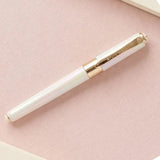 A white iridescent rollerball pen with gold trim and a gold clip, resting diagonally on a textured light pink surface.