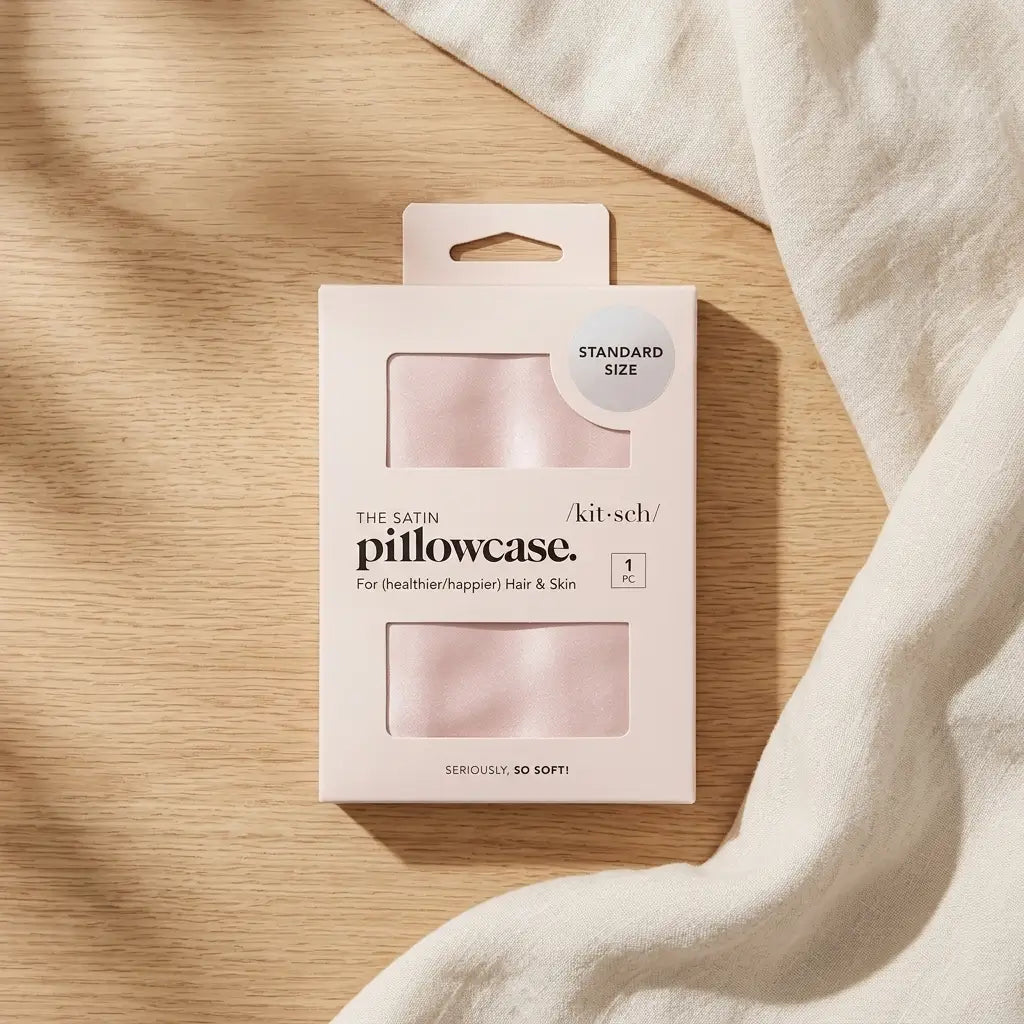 Satin pillowcase in blush pink laid flat beside a folded cloth, a standard size zippered pillowcase that is gentle on hair and skin.