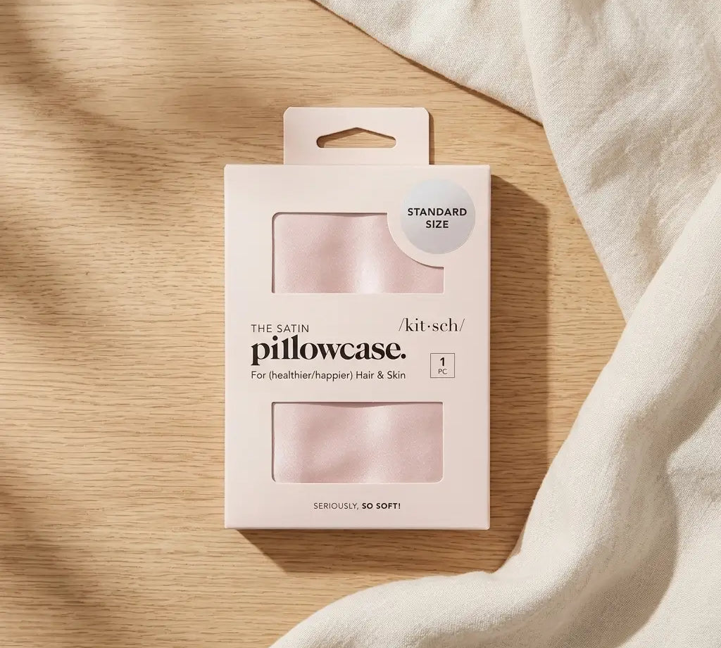 Satin pillowcase in blush pink laid flat beside a folded cloth, a standard size zippered pillowcase that is gentle on hair and skin.