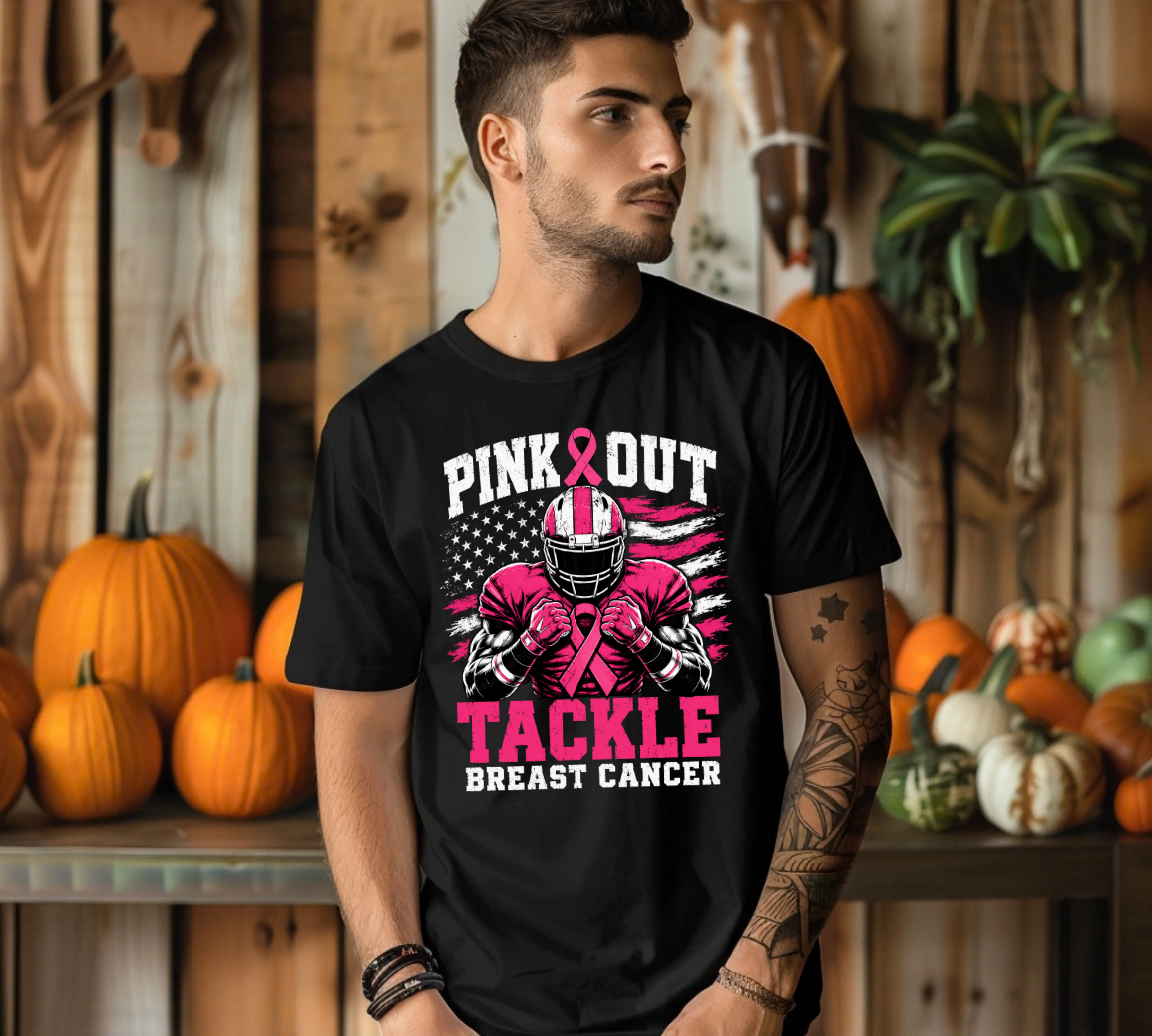A man wearing a black graphic t-shirt with a pink and white 'Pink Out Tackle Breast Cancer' football player design, posed in front of a rustic wooden wall decorated with pumpkins.