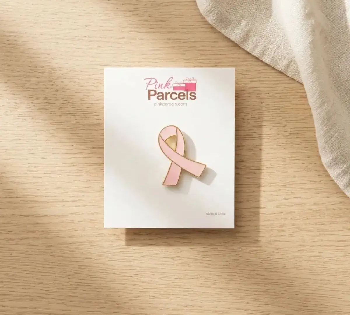 A pink enamel ribbon pin by Pink Parcels, displayed on its white backing card, set on a light wood tabletop with a draped fabric in the corner.