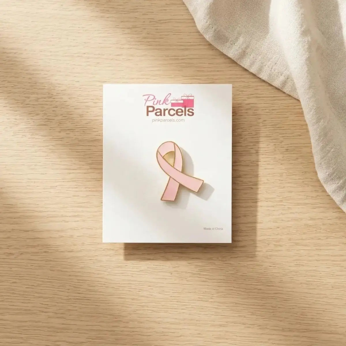 A pink enamel ribbon pin by Pink Parcels, displayed on its white backing card, set on a light wood tabletop with a draped fabric in the corner.