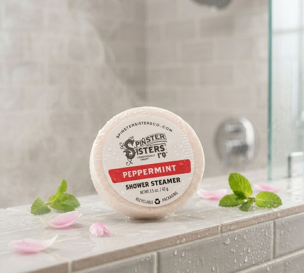 Peppermint shower steamer tablet resting on a tiled shower ledge with water droplets, pink petals, and fresh mint leaves.