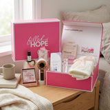 The Pink Signature Box open on a wooden bedside table, holding a handheld fan, peppermint oil, satin pillowcase, shower steamer, ivory scarf, and journal.