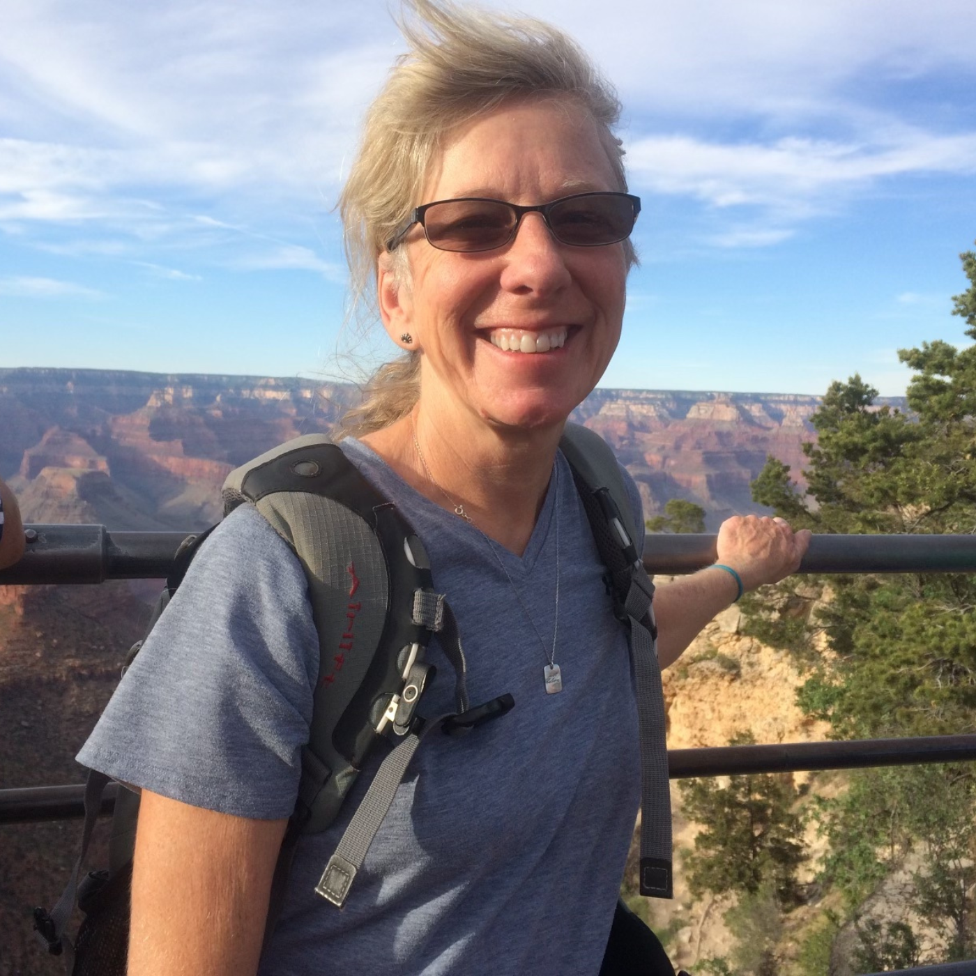 Theresa Strong, a smiling woman with  blonde hair and sunglasses stands outdoors at a canyon overlook, wearing a gray t-shirt and a hiking backpack, with the Grand Canyon landscape and blue sky in the background.