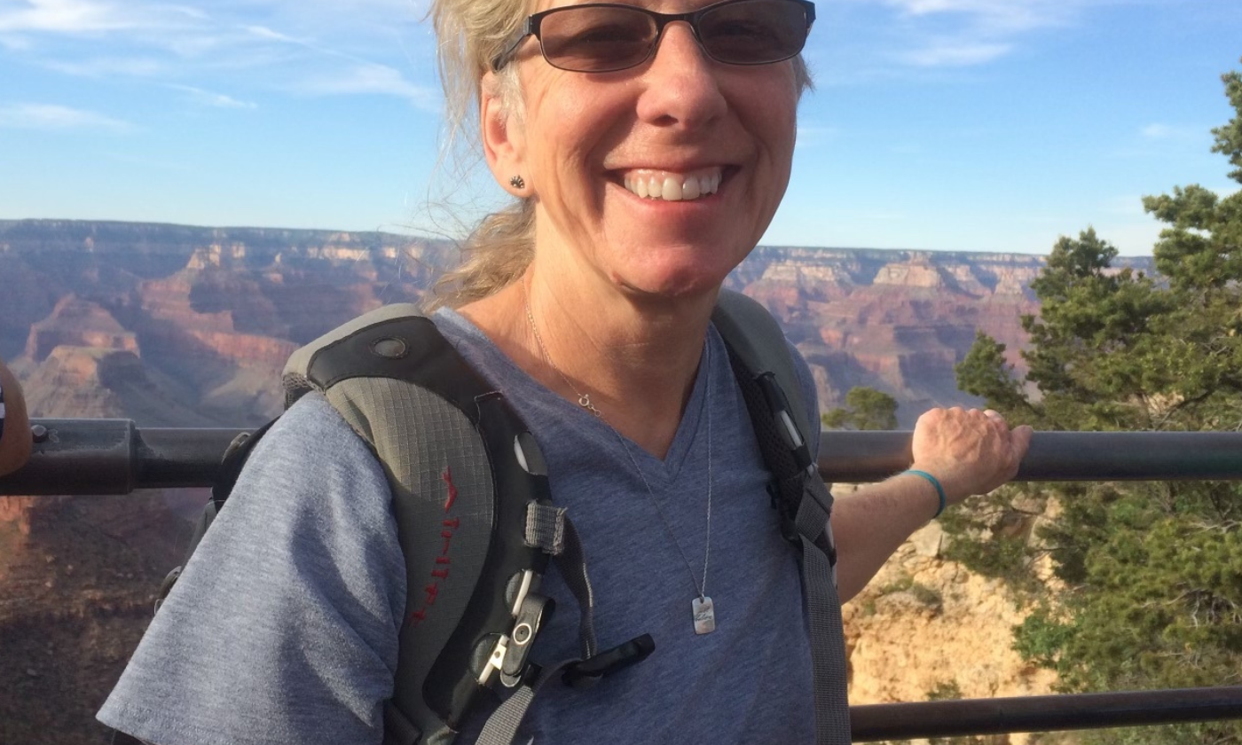 Theresa Strong, a smiling woman with  blonde hair and sunglasses stands outdoors at a canyon overlook, wearing a gray t-shirt and a hiking backpack, with the Grand Canyon landscape and blue sky in the background.