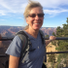 Theresa Strong, a smiling woman with  blonde hair and sunglasses stands outdoors at a canyon overlook, wearing a gray t-shirt and a hiking backpack, with the Grand Canyon landscape and blue sky in the background.