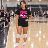 A young woman stands on an indoor volleyball court wearing a black graphic T-shirt that reads 'PINK OUT Together We Fight' in pink and white, paired with black shorts, white knee pads, white socks, and pink sneakers.