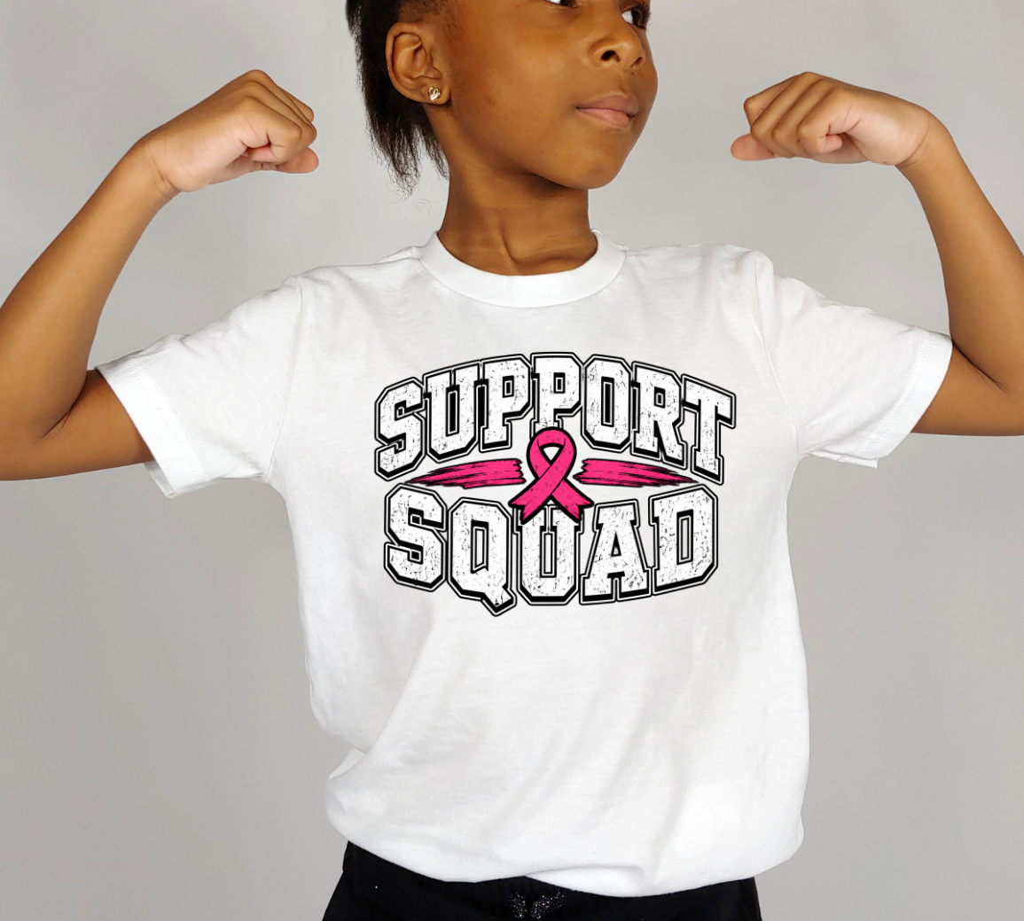 A child wearing a white graphic T-shirt with the words 'SUPPORT SQUAD' and a pink ribbon across the chest, posed with arms flexed against a plain light gray background.