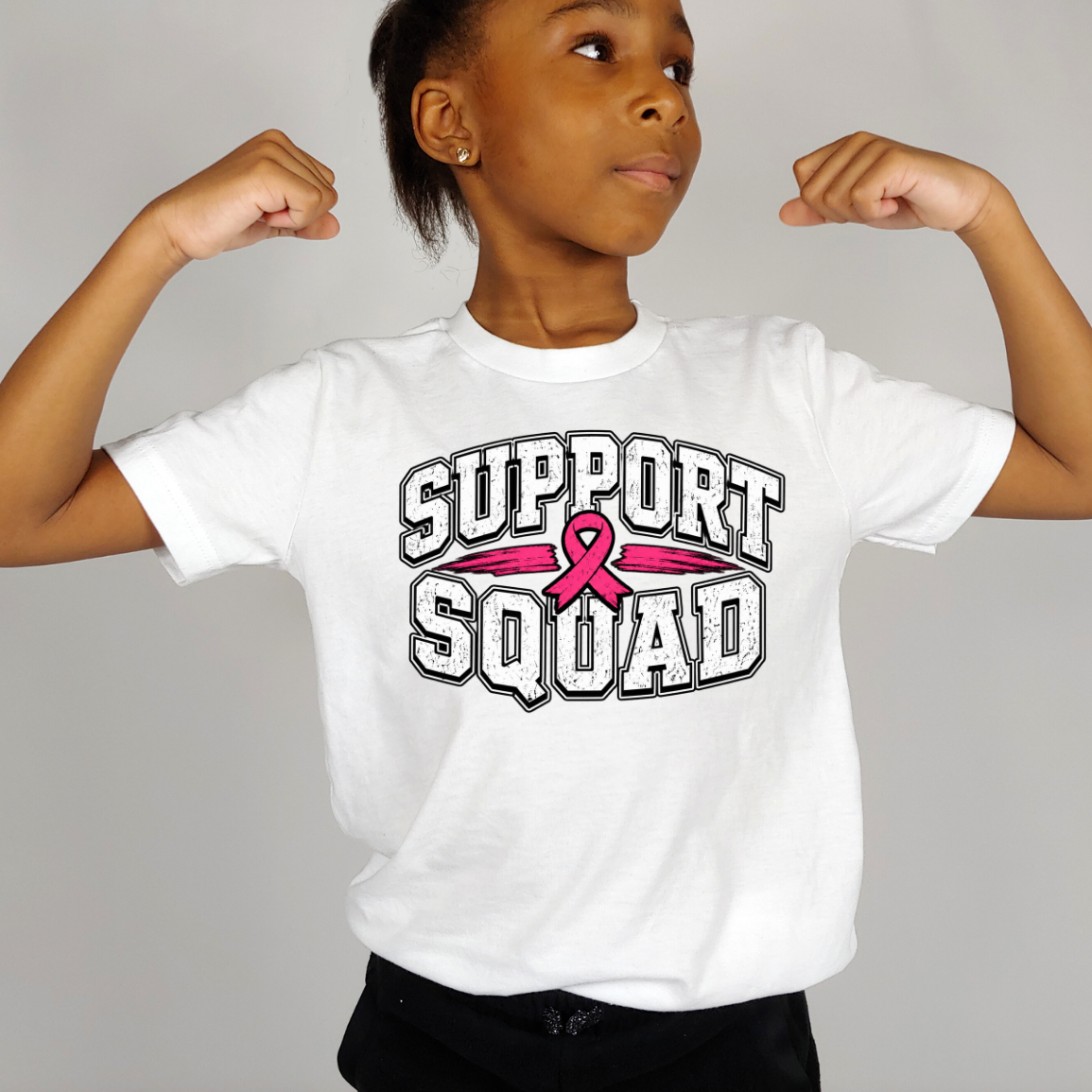 A child wearing a white graphic T-shirt with the words 'SUPPORT SQUAD' and a pink ribbon across the chest, posed with arms flexed against a plain light gray background.